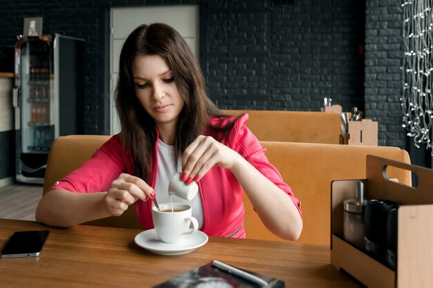 A young girl is pouring cream or milk into coffee in a cafe on wooden table