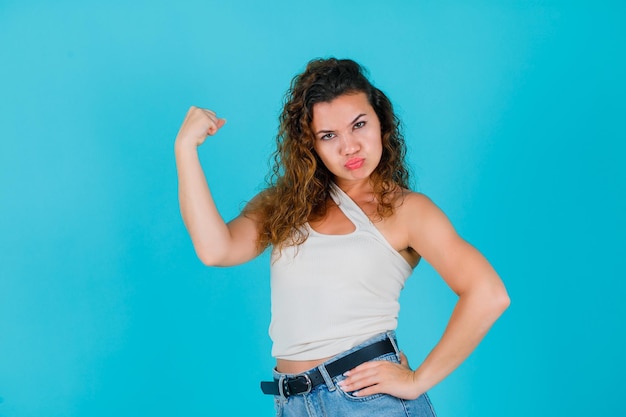 Фото Молодая девушка показывает свои мышцы и кладет другую руку на талию на синем фоне