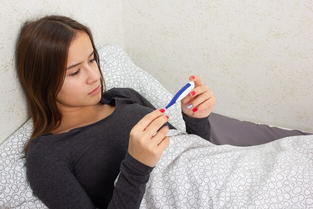 A young girl is sick at home in bed, holds a thermometer.