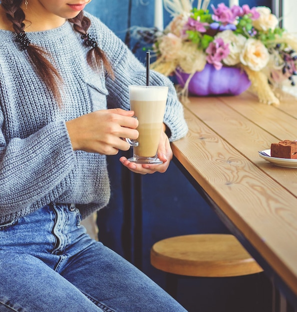 Photo a young girl is sitting in a cafe and holding a cup of coffee in her hands. beautiful glass mug with a straw.