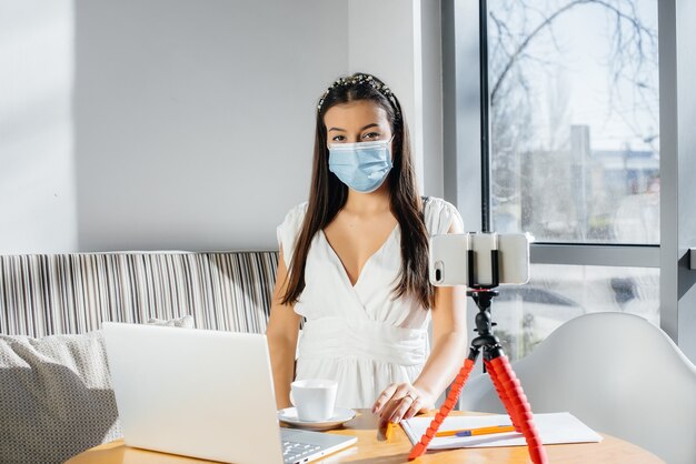 Una giovane ragazza è seduta in un caffè con una maschera e dirige un video blog. comunicazione alla telecamera.