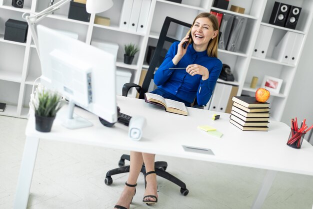 Foto una ragazza è seduta alla scrivania di un computer, con una matita in mano e parla al telefono.