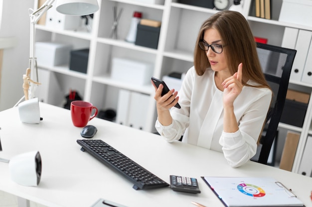 A young girl is sitting at the desk in the office and is holding a phone.