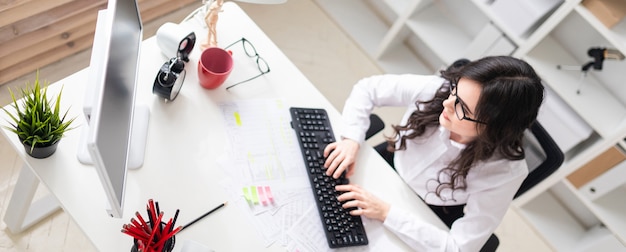 A young girl is sitting in the office at the computer and working with documents.