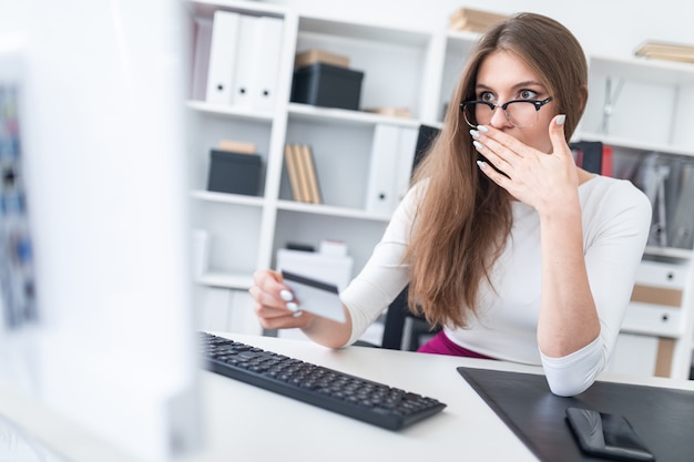 A young girl is sitting at a table, looking at a Bank card and covered her mouth with her hand.