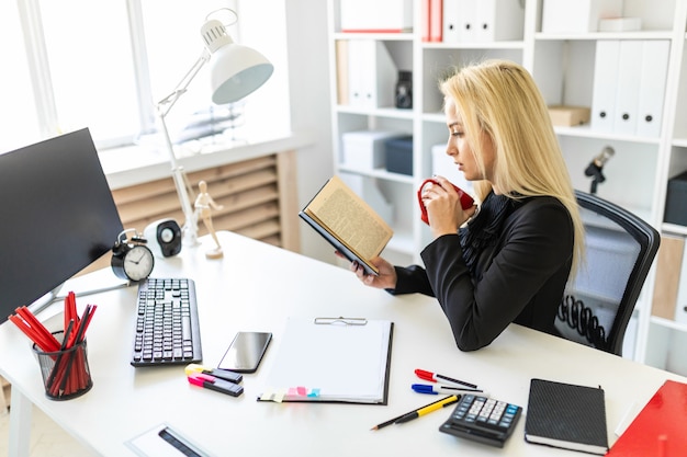 A young girl is sitting at a table in the office, holding a cup and reading a book.