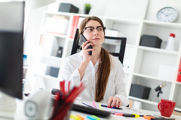 Una ragazza è seduta a un tavolo in ufficio e tiene in mano una penna e parla al telefono.