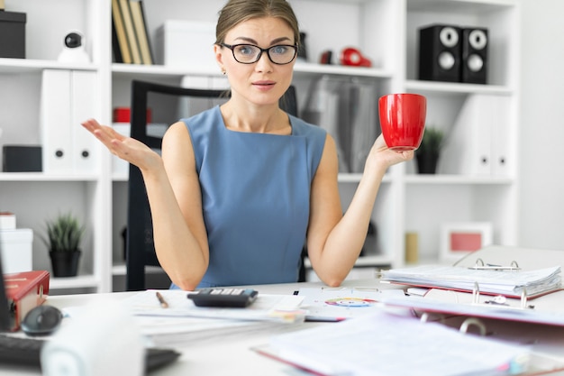 A young girl is sitting at a table in the office, holding a red cup in her hand and working with documents.