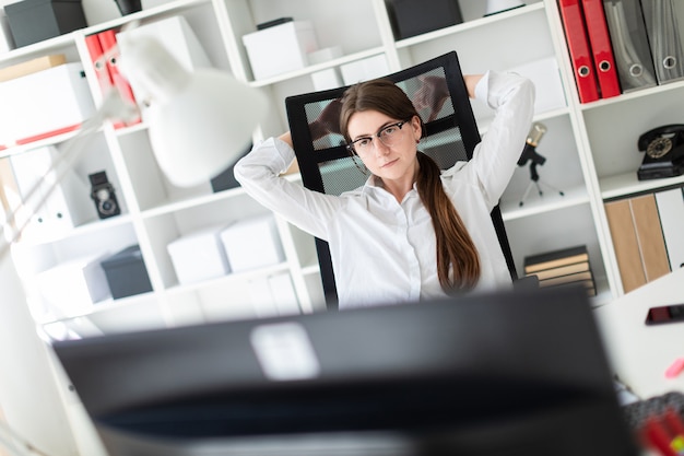 A young girl is sitting at a table in the office, laying her hands behind her head and looking at the monitor.