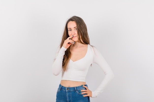 Young girl is smiling by holding forefinger on lips and other hand on waist on white background