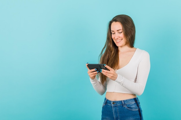 Young girl is smiling by looking at smartphone screen on blue background