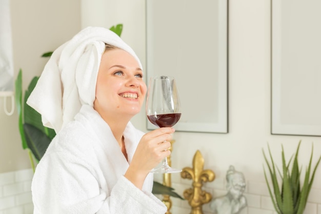 молодая девушка улыбается в белом халате и банном полотенце на голове, держа в руках стеклянный стакан красного