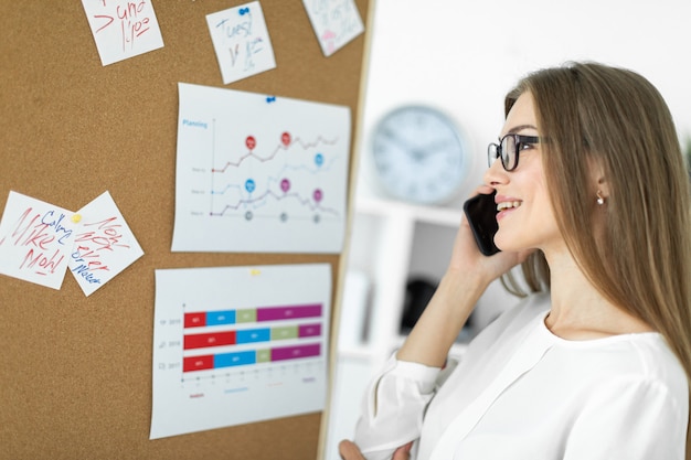 A young girl is standing near the board with stickers and talking on the phone.