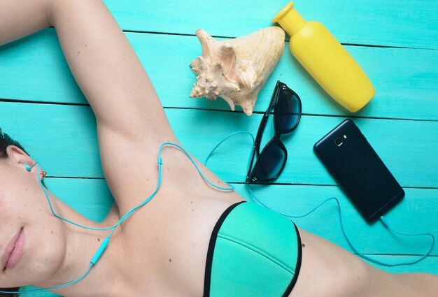 A young girl is sunbathing and listening to music through headphones. Accessories for the resort rest on the beach: sunblock, sunglasses, shell. Top view.