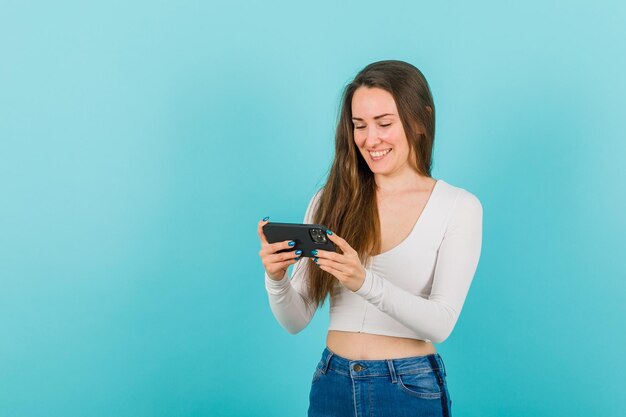 Young girl is taking photo with smartphone by smiling on blue background