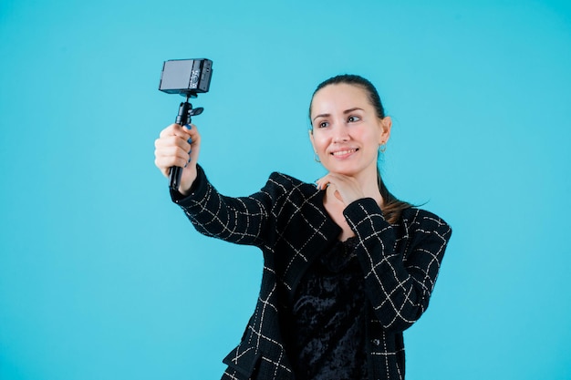 Young girl is taking selfie with her camera by putting hand under chin on blue background