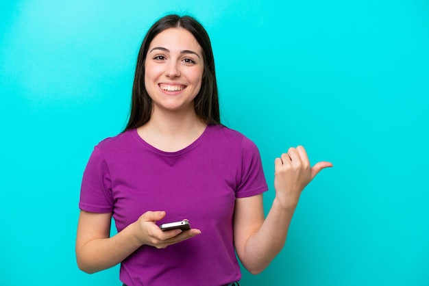 Молодая девушка изолирована на синем фоне с помощью мобильного телефона и указывая на боковую сторону