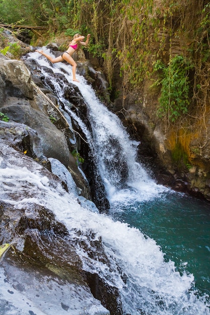 Young girl jumping from high rock to natural water pool under waterfall in tropical mountains