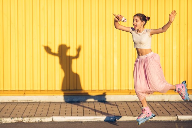 Young girl jumping on roller skates and eating ice cream on yellow wall