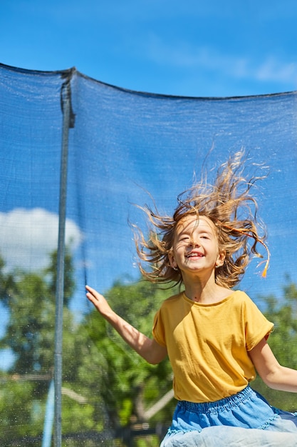 A young girl jumping up and down on her trampoline outdoors, in the backyard of the house on a sunny summer day, summertime vacation.