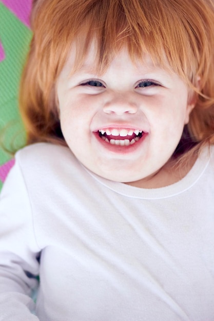 Young girl laughing and funny portrait of a baby on a home playpen ground with a smile Ginger infant kid laugh and happy in a house with joy youth and positivity from childhood looking up