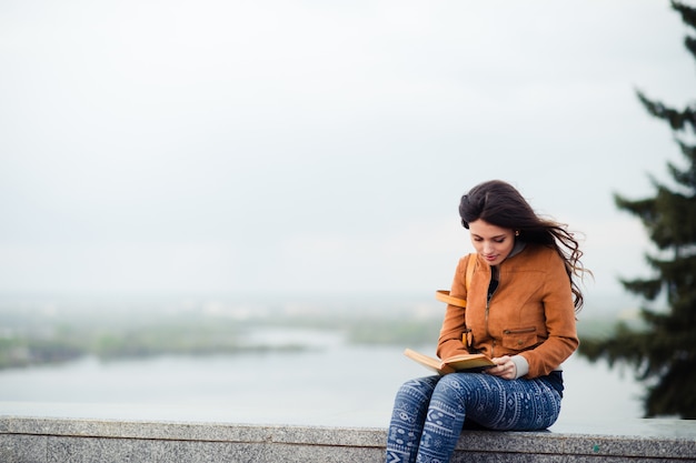 Молодая девушка в кожаном плаще сидит с книгой на берегу реки в прохладный осенний весенний день