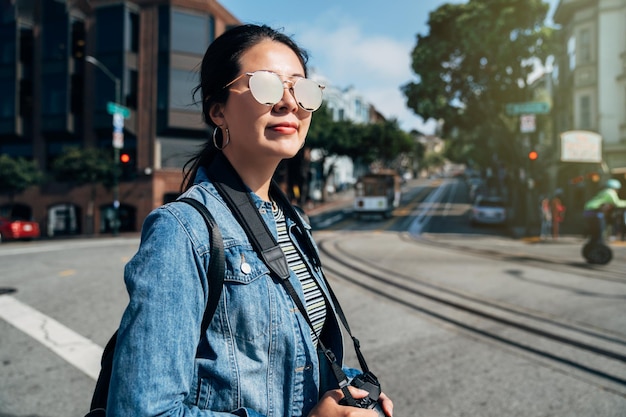 Giovane ragazza lente uomo che cammina sulla trafficata strada urbana di san francisco in una giornata di sole tenendo la fotocamera reflex. metropolitana leggera sullo sfondo. bella signora che indossa occhiali da sole visita la città in america.