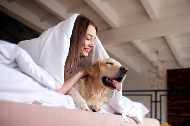 사진 어린 소녀는 골든 리트리버 개와 함께 따뜻한 담요 아래 침대에 누워 창밖을 내다봅니다.