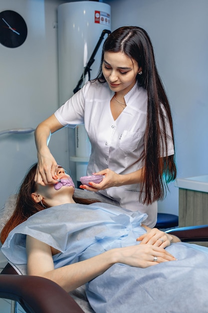 Young girl lies with patches under her eyes in chair in spa salon for procedure to eliminate flaws on her face. Beautician makes cream mask.