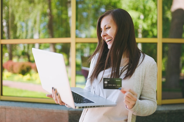 Ragazza in abiti casual leggeri in possesso di carta di credito. donna che lavora al computer portatile moderno vicino all'edificio a specchio con riflesso dell'albero in strada all'aperto. ufficio mobile. concetto di business freelance.