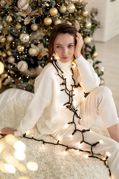 クリスマスツリーの背景にライトな服を着た若い女の子がベッドに座って新年を楽しんでいます