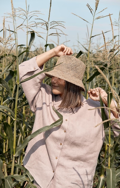여름 화창한 날 린넨 양복과 모자를 쓴 어린 소녀는 여행을 좋아하는 옥수수 밭에 서 있습니다.