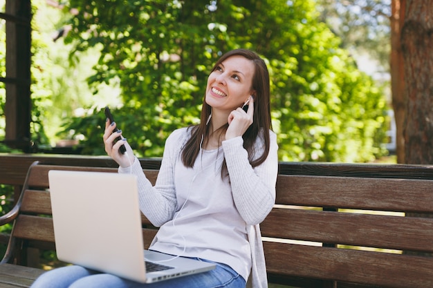 Young girl listen music in mobile phone with earphones. Woman sitting on bench working on modern laptop pc computer in city park in street outdoors on nature. Mobile Office. Freelance business concept