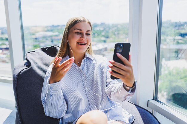 A young girl listens to music on headphones and watches something on a mobile phone Modern woman lifestyle concept Young woman sitting in coworking space office interior