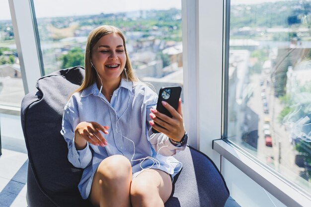 A young girl listens to music on headphones and watches something on a mobile phone Modern woman lifestyle concept Young woman sitting in coworking space office interior