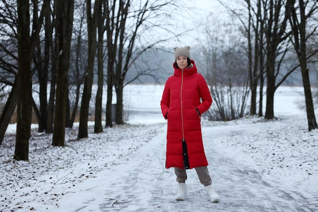 A young girl in a long red down-padded coat stands in the winter forest.