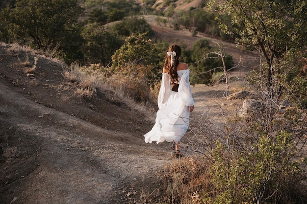 A young girl in a long white dress is walking down a path in a hilly area among trees The concept of freedom and travel