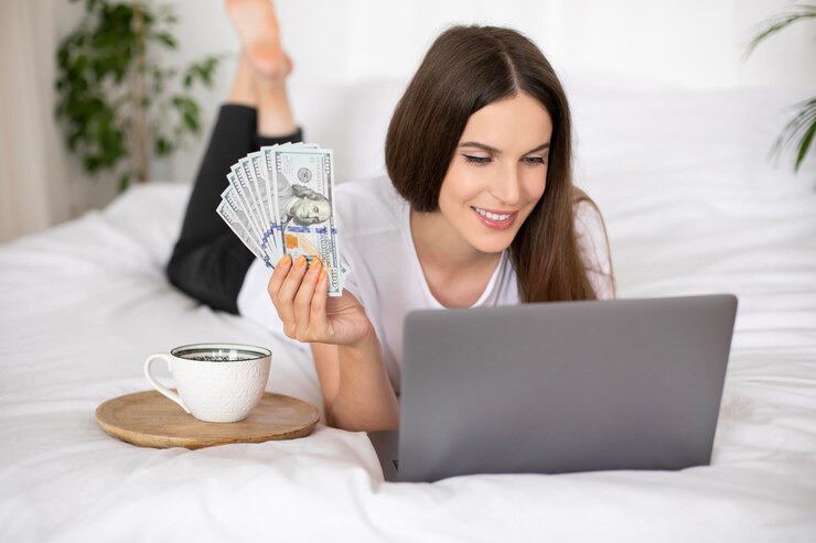 ベッドに横になってドル通貨を保持している若い女の子は、自宅でラップトップコンピューターの仕事を使用しています