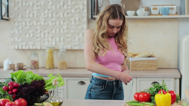 Молодая девушка измеряет свою талию на кухне возле стола с овощами и фруктами Концепция здорового питания и диеты