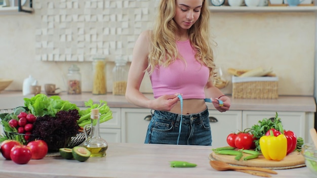 Young girl measures her waist in the kitchen near the table with vegetables and fruits Concept of healthy eating and diet