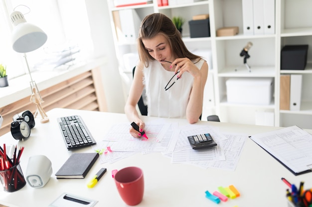 A young girl in the office is holding a pink marker, glasses and working with the documentation.