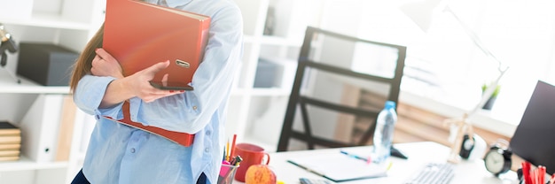 A young girl in the office is standing, leaning on a table, and is holding a phone and a folder with documents.