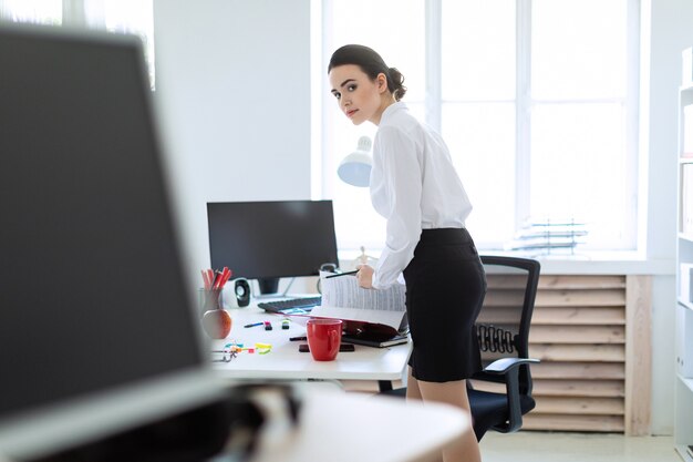 A young girl in the office is standing near the table, holding a pencil in her hand and leafing through the folder with the documents.