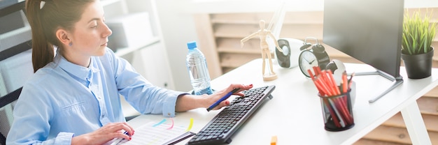 A young girl in the office sits at a table, holds a pen in her hand and works at the computer.