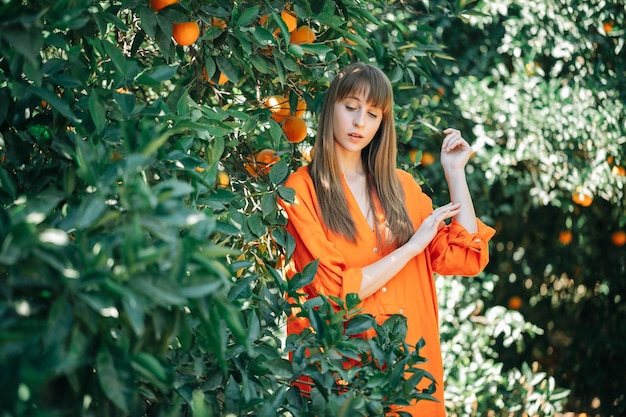 Giovane ragazza in abito arancione sta guardando in basso alzando le mani nel giardino degli aranci