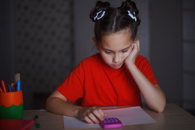 A young girl in an orange T-shirt is studying sitting at a desk. Counts the numbers on the calculator.