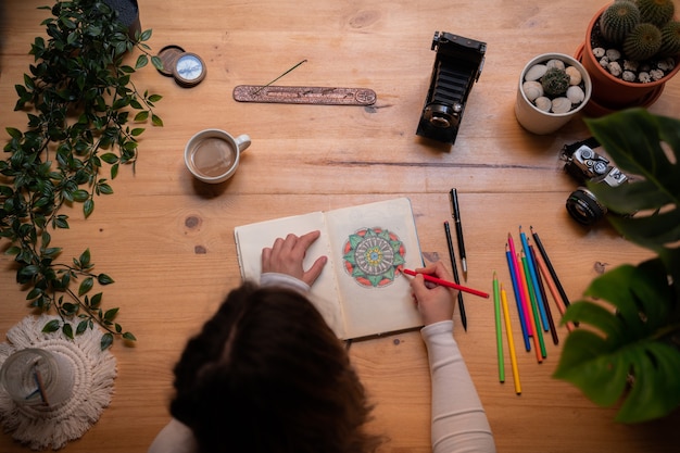 Una giovane ragazza che dipinge un mandala con matite di molti colori, su un tavolo di legno. ha pennelli, incenso, macchine fotografiche e una grande finestra. vista dall'alto.