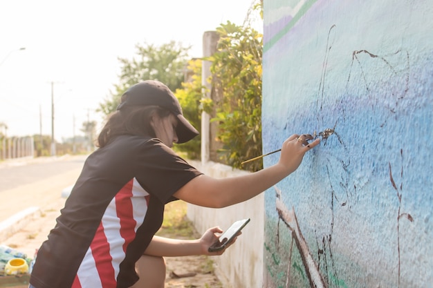 携帯電話を参考にして街の壁を描いている少女。