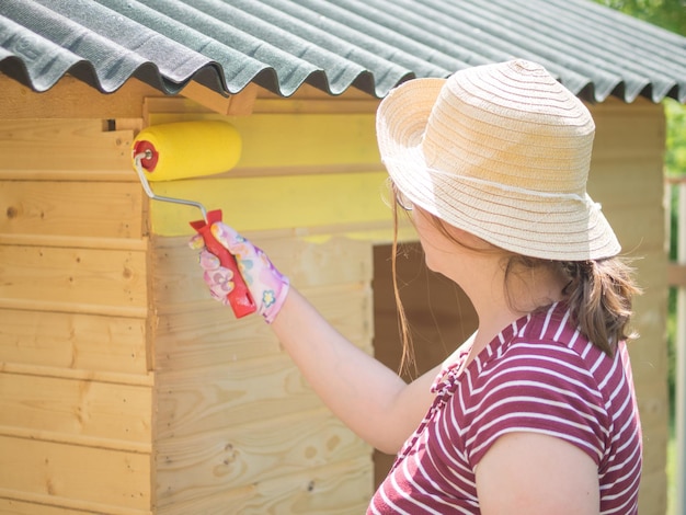 Young girl paints a wooden wall with a roller with yellow paint