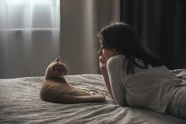 a young girl in pajamas lies on the bed near the window next to her domestic cat